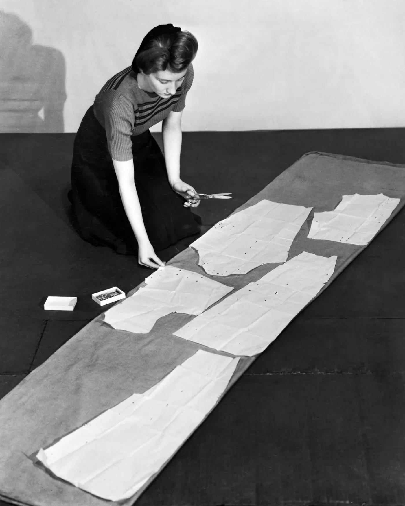 A pattern cutter kneeling over paper pattern pieces laid along a length of cloth.